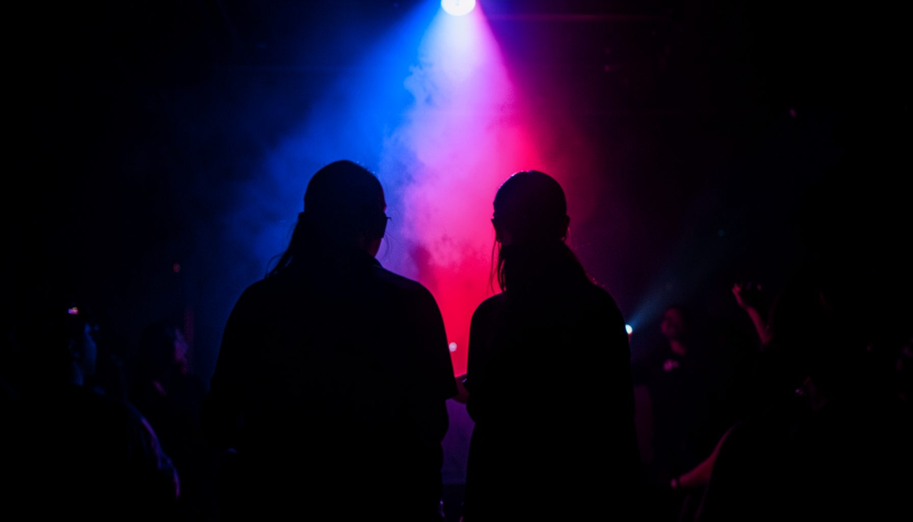 Dos siluetas bajo un foco azul y rojo sobre un fondo oscuro, atmósfera de club nocturno, tonos profundos y eléctricos