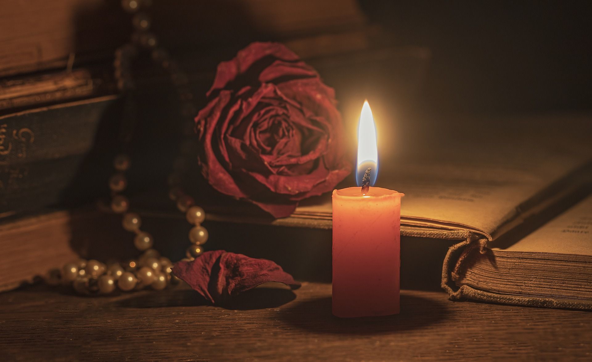 Rincón de lectura con una vela encendida junto a una rosa roja seca, libros antiguos y perlas, en una atmósfera cálida y tenue
