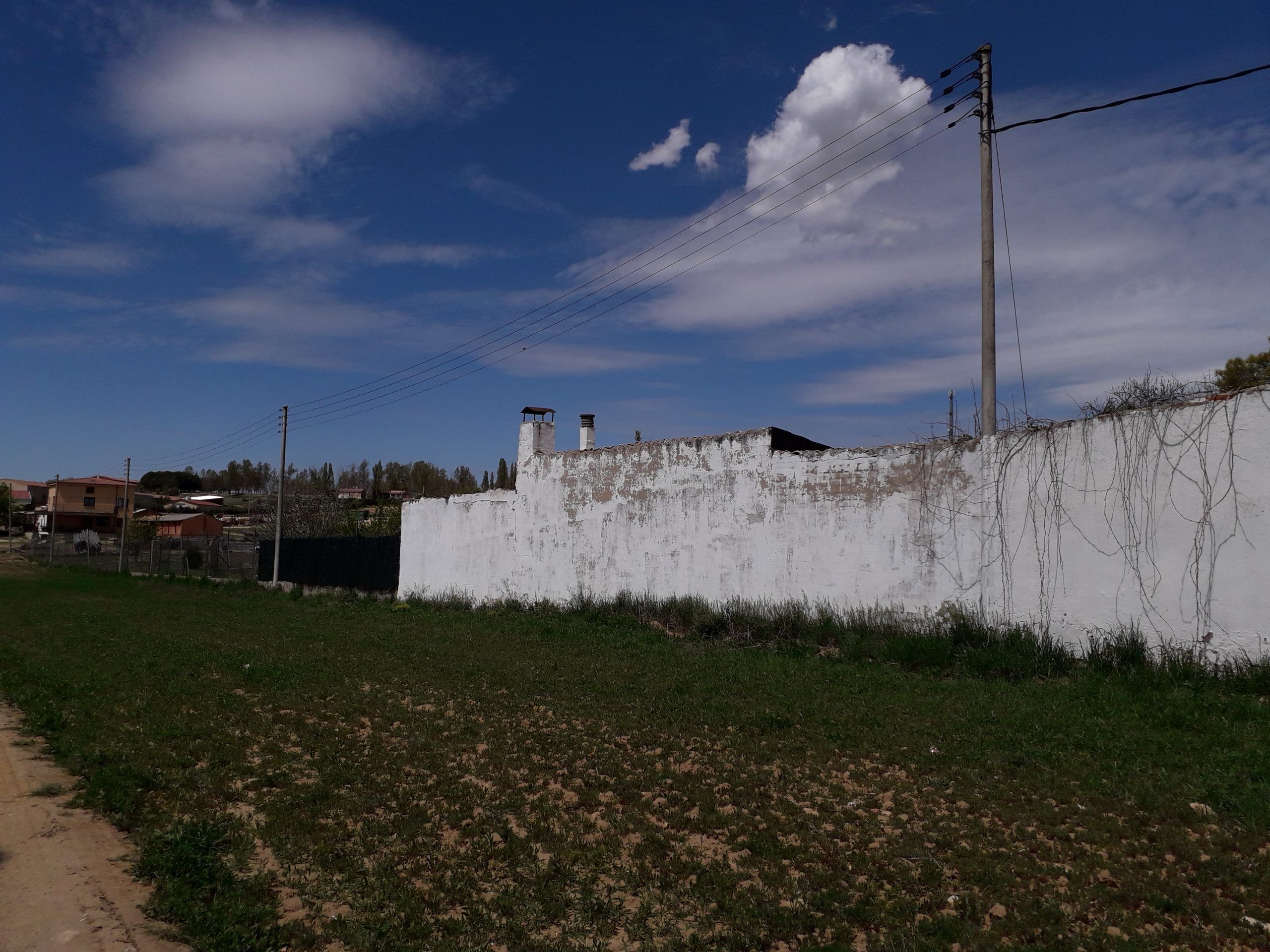 Muro encalado junto a un campo de hierba, con postes eléctricos y edificios lejanos bajo cielo azul