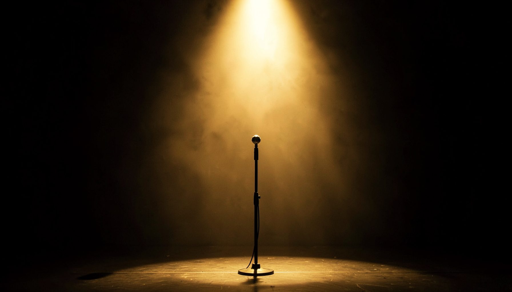 Luz dorada cenital sobre un escenario vacío con un micrófono de pie, atmósfera solemne y teatral, tonos ámbar y sombra